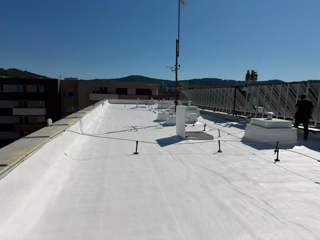 Finální hydroizolace ploché střechy s fotovoltaikou chráněná bílým silikonovým nátěrem proti UV záření. Realizace Jínce.